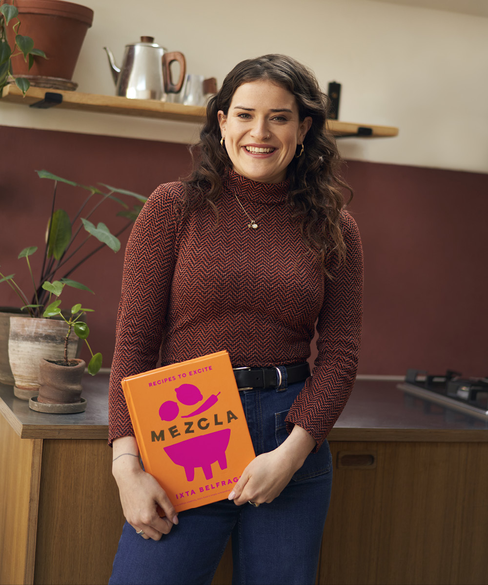 Ixta Belfrage stands in a kitchen holding a copy of her book, Mezcla